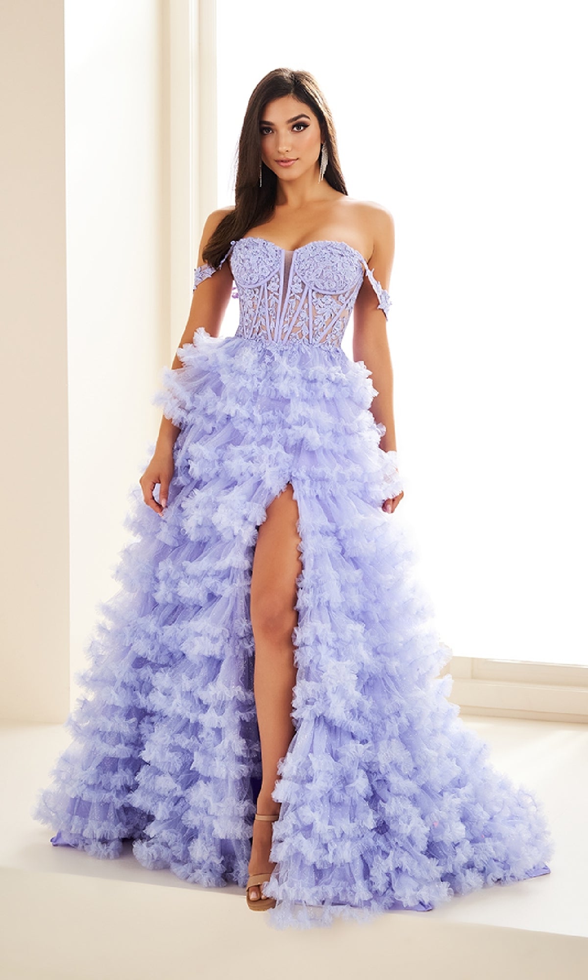 A woman poses gracefully indoors by a bright window wearing the Ellie Wilde EW36201, a lavender off-the-shoulder long prom dress with a ruffled skirt, lace applique corset bodice, and a high slit.